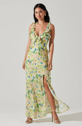 The dress is a sleeveless, ankle-length summer gown with shoulder straps tied into bows. It features a deep V neckline with ruffles along the front. The fabric has a colorful, abstract floral pattern in shades of green, blue, yellow, and orange. The dress includes a high slit on one side, revealing the leg, and the material appears lightweight and flowy.